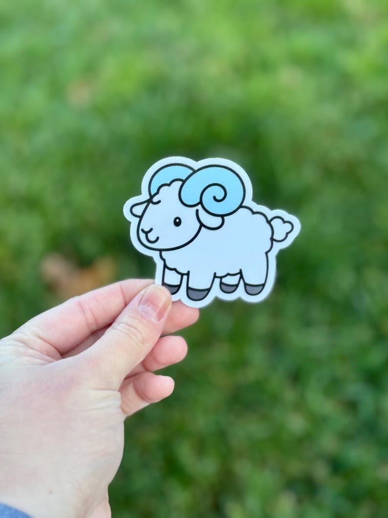 May include: A sticker depicting a cartoon ram with light blue fur and curly horns. The ram has black hooves and a smiling face. The sticker is on a white background.