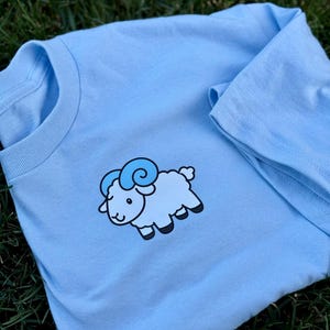 May include: A light blue t-shirt featuring a cartoon ram design. The ram is light blue with darker blue horns and black hooves. The shirt is made of soft material and is suitable for casual wear.