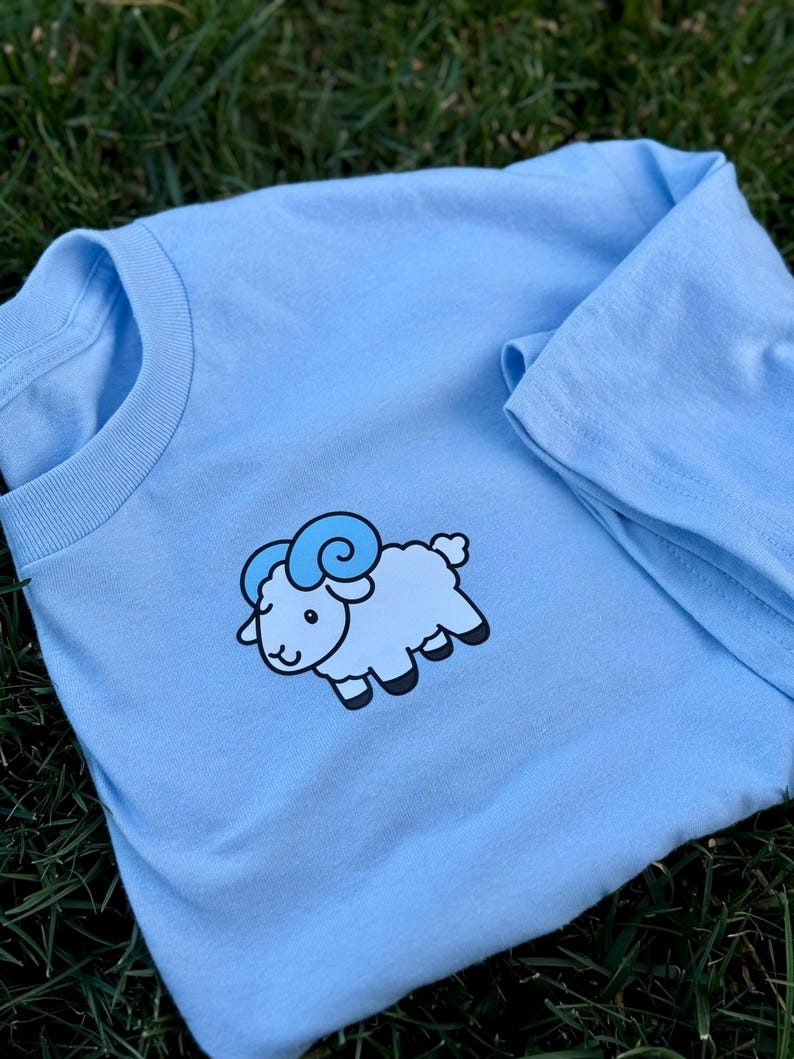 May include: A light blue t-shirt featuring a cartoon ram design. The ram is light blue with darker blue horns and black hooves. The shirt is made of soft material and is suitable for casual wear.