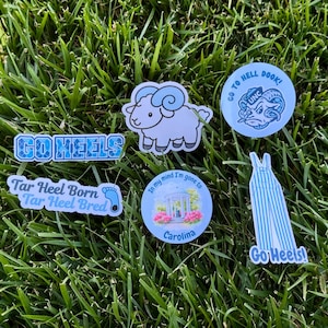 Puede incluir: Una colecci&oacute;n de pegatinas y pines con tem&aacute;tica de Carolina, con un carnero, las palabras "Go Heels" y otras frases relacionadas. Los art&iacute;culos son en tonos de azul y blanco, sobre un fondo de hierba verde.