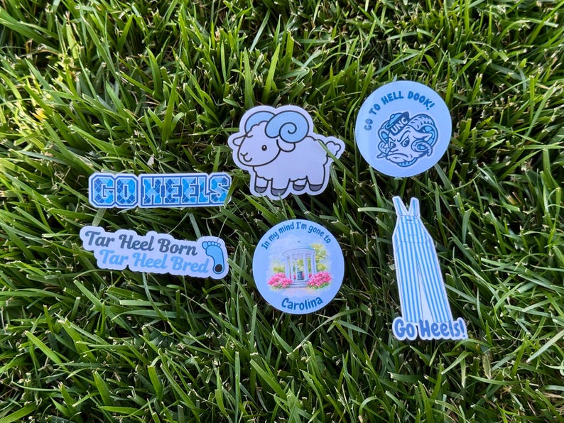 Puede incluir: Una colecci&oacute;n de pegatinas y pines con tem&aacute;tica de Carolina, con un carnero, las palabras "Go Heels" y otras frases relacionadas. Los art&iacute;culos son en tonos de azul y blanco, sobre un fondo de hierba verde.