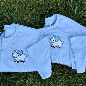May include: Two light blue long-sleeved shirts with a cartoon ram graphic on the front. The ram has a white body, black hooves, and blue horns. The shirts are displayed on a bed of green grass.