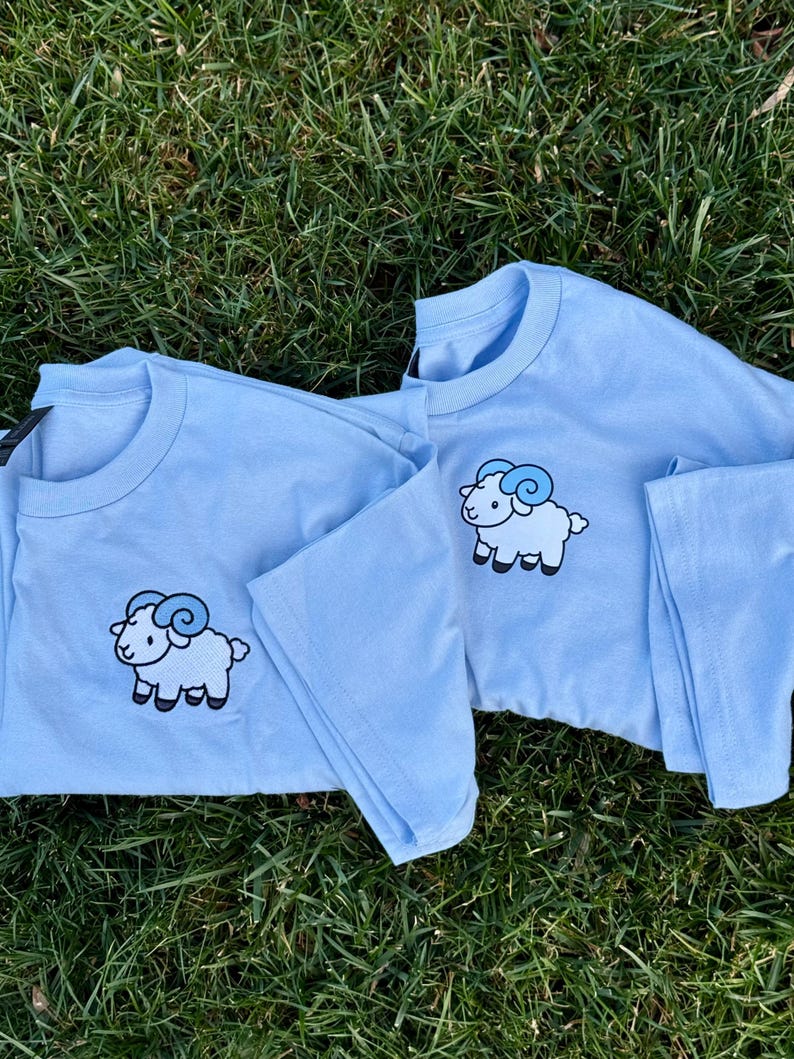May include: Two light blue long-sleeved shirts with a cartoon ram graphic on the front. The ram has a white body, black hooves, and blue horns. The shirts are displayed on a bed of green grass.