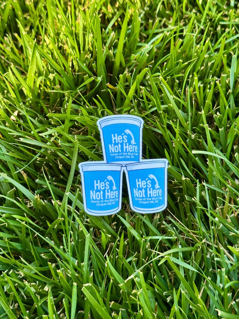Puede incluir: Tres pegatinas con un dise&ntilde;o azul y blanco, con el texto "He's Not Here" y "Home of the Blue Cup, Chapel Hill, NC". Las pegatinas est&aacute;n colocadas sobre un lecho de hierba verde.