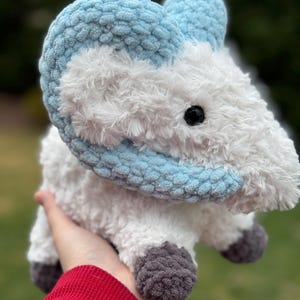 May include: A handmade plush ram toy with a fluffy white body and light blue horns. The ram has gray hooves and a black eye. The toy is made of soft, textured yarn, perfect for a nursery or as a decorative item.