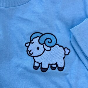 May include: Light blue long-sleeved shirt featuring an embroidered cartoon ram. The ram has light blue horns and a light blue body with black hooves. The shirt's neckline and sleeve are visible.
