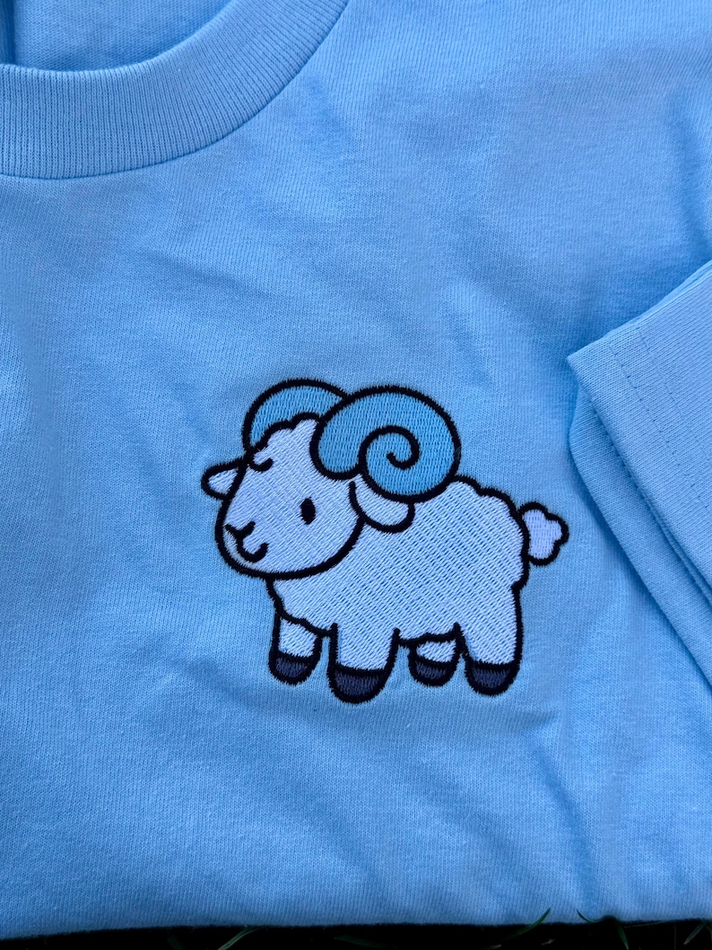 May include: Light blue long-sleeved shirt featuring an embroidered cartoon ram. The ram has light blue horns and a light blue body with black hooves. The shirt's neckline and sleeve are visible.