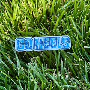 Puede incluir: Una pegatina rectangular con el texto "GO HEELS" en azul y blanco. Las letras tienen un patr&oacute;n de diamantes. La pegatina est&aacute; colocada sobre un fondo de hierba verde.