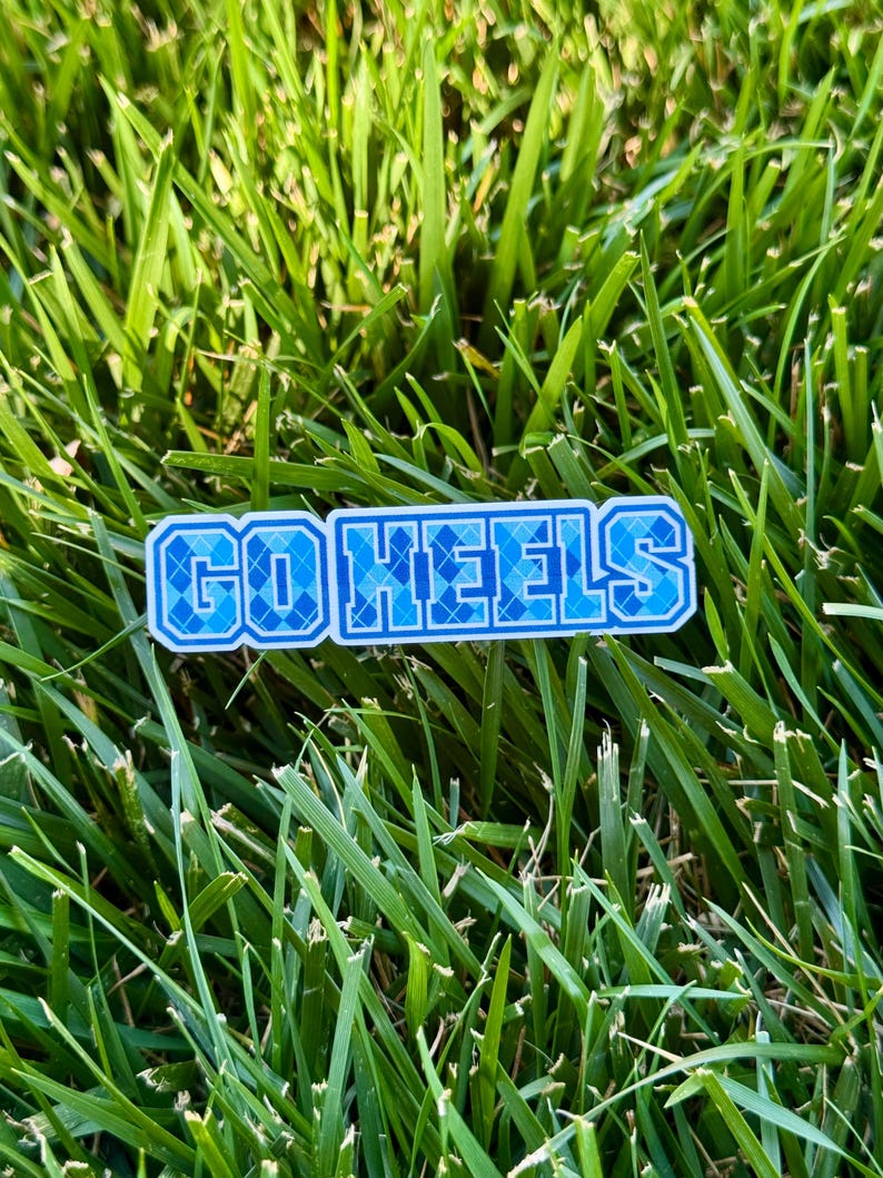 Puede incluir: Una pegatina rectangular con el texto "GO HEELS" en azul y blanco. Las letras tienen un patr&oacute;n de diamantes. La pegatina est&aacute; colocada sobre un fondo de hierba verde.