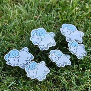 May include: Seven cartoon sheep stickers are scattered on green grass. The sheep are white with light blue horns and a glitter effect. The stickers are designed with a cartoon style, featuring a cute and friendly appearance.