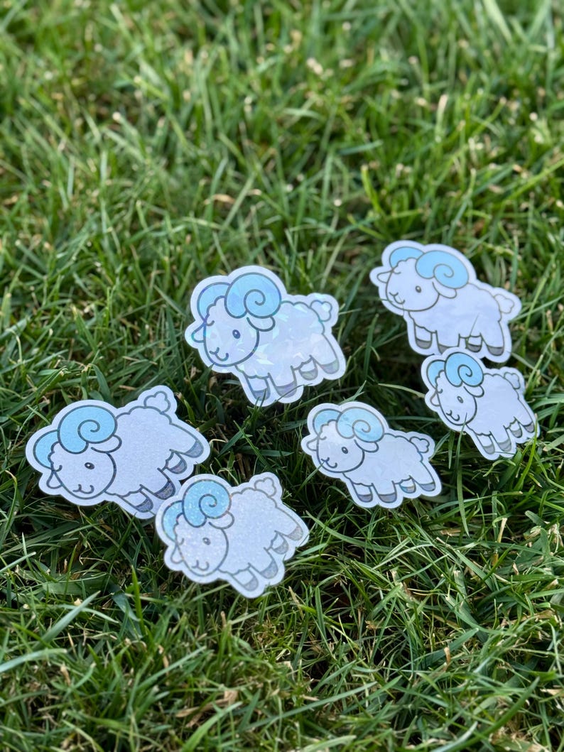 May include: Seven cartoon sheep stickers are scattered on green grass. The sheep are white with light blue horns and a glitter effect. The stickers are designed with a cartoon style, featuring a cute and friendly appearance.