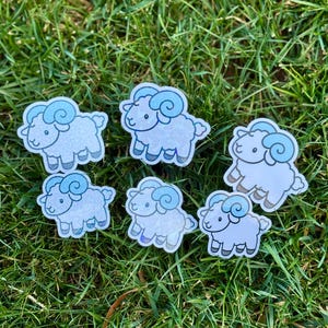 May include: Six stickers featuring a cartoon ram with light blue horns and accents. The stickers are white with a subtle glitter effect and are arranged on green grass.
