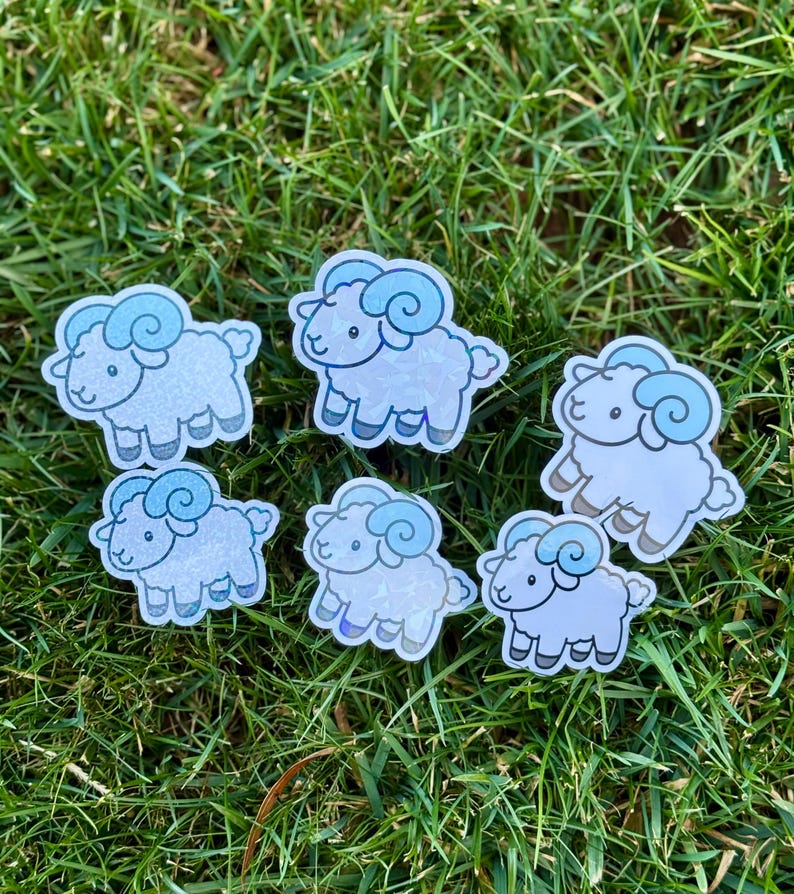 May include: Six stickers featuring a cartoon ram with light blue horns and accents. The stickers are white with a subtle glitter effect and are arranged on green grass.