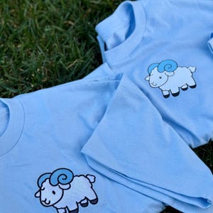 May include: Two light blue t-shirts with a cartoon ram graphic. The ram is white with blue accents and black hooves. The shirts are laying on green grass.