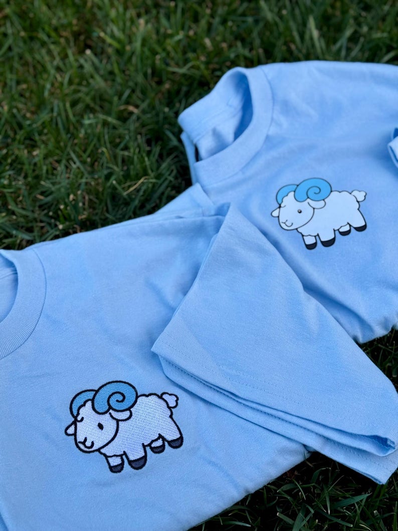 May include: Two light blue t-shirts with a cartoon ram graphic. The ram is white with blue accents and black hooves. The shirts are laying on green grass.
