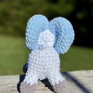May include: A handmade crochet elephant toy, viewed from the back, with light blue ears, a white body, and gray feet. The plush toy is sitting on a wooden surface with a blurred green background.