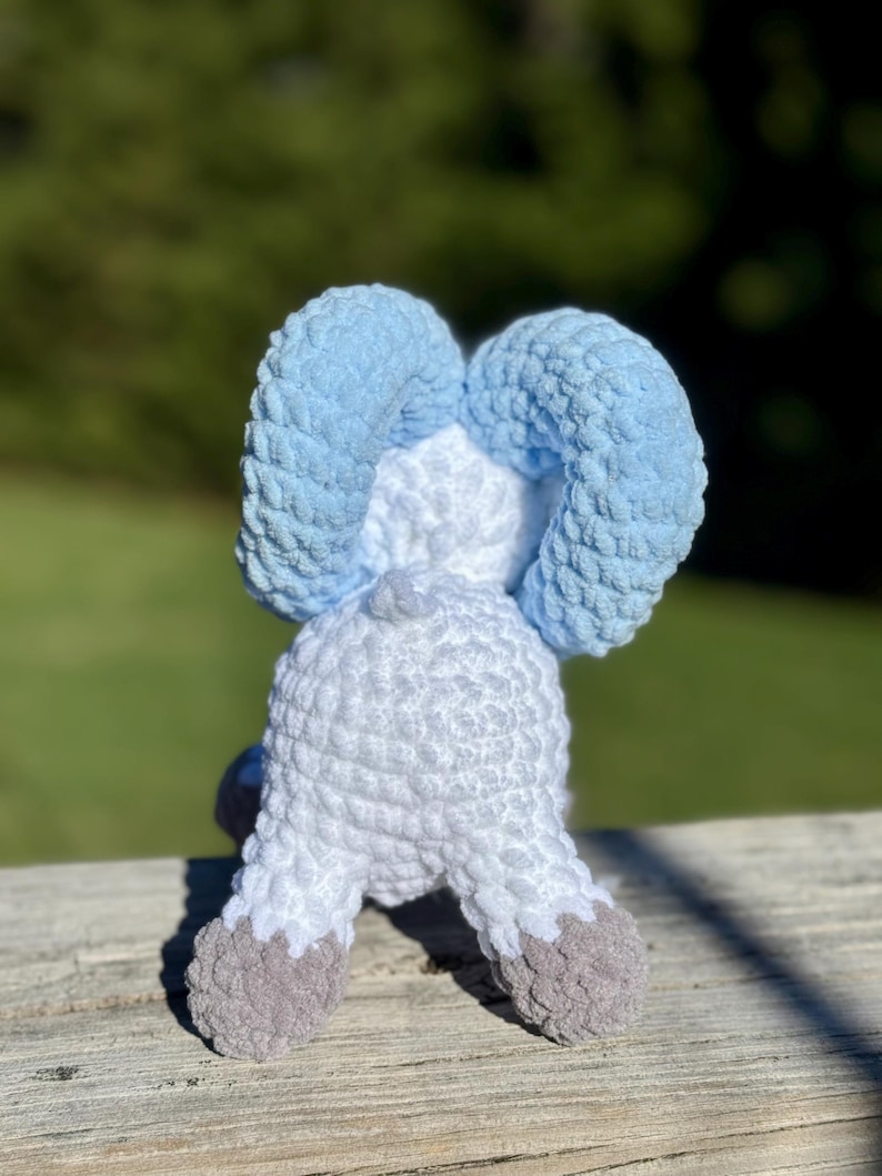 May include: A handmade crochet elephant toy, viewed from the back, with light blue ears, a white body, and gray feet. The plush toy is sitting on a wooden surface with a blurred green background.