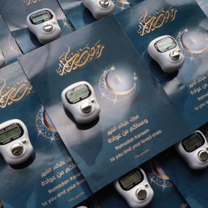 May include: An overhead shot shows several small, white digital tally counters with a silver button and a small screen. They are arranged on blue cards with gold Arabic script and a crescent moon design. The cards have text in English and Arabic.