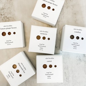 May include: Six white boxes with brown soap bars inside. Each box has a different scent: lemon ginger honey, turmeric, comfrey, chamomile, carrot. The boxes are labeled with the scent and "sabu brooklyn" and "soap bar hand poured vegan 5.0 oz."