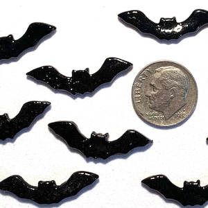 May include: A collection of ten black bat-shaped embellishments, some with glitter, arranged around a dime. The bats have a glossy finish and are spread out on a white surface. Ideal for Halloween crafts and decorations.