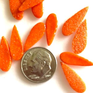 May include: Close-up of several small, orange, teardrop-shaped beads with a textured surface. A silver dime is included for size comparison. The beads are scattered on a white surface.
