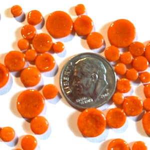 Op de afbeelding: Een verzameling van oranje glazen cirkels in verschillende maten, verspreid over een wit oppervlak. Een zilveren Amerikaanse dime-munt bevindt zich in het midden van de afbeelding als maatstaf.
