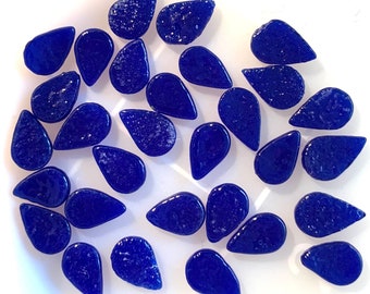 May include: A collection of deep blue, teardrop-shaped glass beads. The beads have a glossy finish and vary slightly in size and texture. They are scattered on a white surface, creating a visually appealing arrangement. Ideal for jewelry making or craft projects.