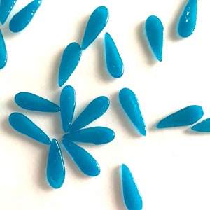 May include: A collection of teal blue glass teardrop shaped beads. The beads are scattered on a white surface.
