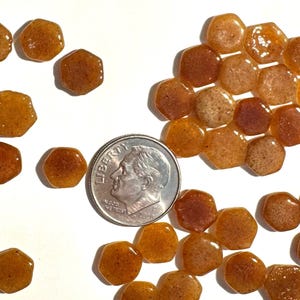 Può includere: Una collezione di piccole tessere esagonali in vetro color ambra con una consistenza maculata. Una moneta da dieci centesimi di dollaro statunitense color argento è posizionata tra le tessere per fornire una scala dimensionale.