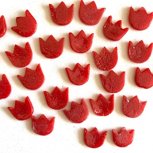 May include: A collection of small, red tulip-shaped glass pieces. The pieces are arranged on a white surface. The glass has a glossy finish, and the shapes are uniform. These decorative pieces could be used for crafts or mosaics.
