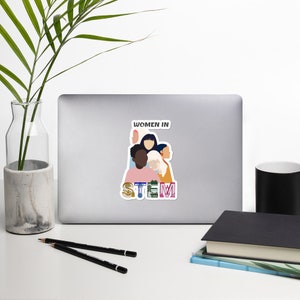 May include: A white sticker on a laptop featuring the text "WOMEN IN STEM" above a graphic of diverse women. The word "STEM" is in colorful letters with science-related imagery.