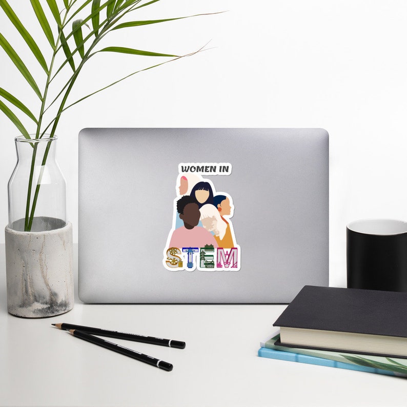 May include: A white sticker on a laptop featuring the text "WOMEN IN STEM" above a graphic of diverse women. The word "STEM" is in colorful letters with science-related imagery.
