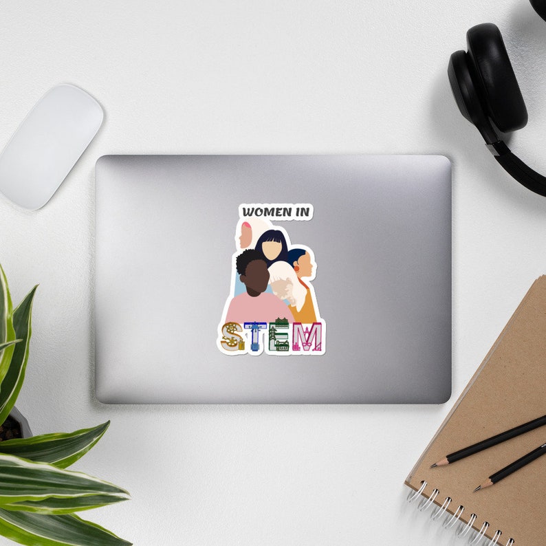 May include: A sticker on a laptop featuring a graphic of diverse women with the text "WOMEN IN STEM." The sticker has a white border and is surrounded by a computer mouse, headphones, and a notebook.