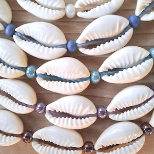 Puede incluir: Un collar de varias hebras con conchas de cauri blancas ensartadas en cordones de colores. Las hebras incorporan pequeñas cuentas redondas y brillantes en tonos azul, morado y marrón, alternándose con las conchas a lo largo del diseño.