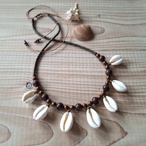 Könnte beinhalten: Eine Halskette mit einer Reihe weißer Kaurimuscheln, die mit dunkelbraunen, kugelförmigen Perlen auf einer dünnen braunen Schnur abwechseln. Das Design umfasst einen langen, verstellbaren Schnurabschnitt mit kleinen dunklen Perlen.