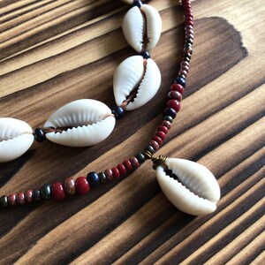 Peut inclure: Gros plan sur un collier composé de coquillages blancs et d'un fil de perles. Les perles sont dans les tons de rouge, marron et bleu foncé. Le collier est posé sur une surface en bois.