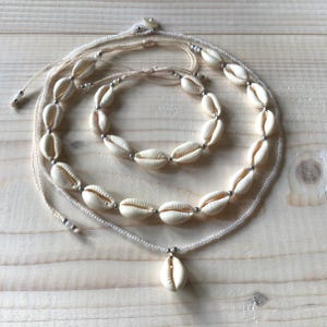 Puede incluir: Un conjunto de tres piezas de joyería de conchas marinas: un collar, una pulsera y un collar con colgante. La joyería presenta conchas marinas de color crema ensartadas con cuentas plateadas y hilo blanquecino.
