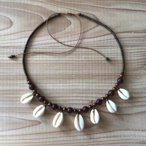 Può includere: Una collana composta da una fila di piccole perline metalliche scure e perline sferiche marroni più grandi. Sei conchiglie di ciprea bianche sono fissate a intervalli lungo la parte inferiore. Include un cordino marrone per la chiusura.