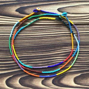 Puede incluir: Tres collares de cuentas de colores. Cada collar está hecho de pequeñas cuentas redondas en una variedad de colores, incluyendo rojo, naranja, amarillo, verde, azul y morado. Los collares están dispuestos en un estilo en capas, con cada hebra ligeramente desplazada.