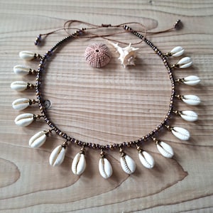 Op de afbeelding: Een ketting met een donkerbruin koord geregen met kleine metalen kralen en witte kaurischelpen als bedels. De ketting heeft verstelbare bruine koordjes aan de bovenkant en wordt getoond naast een kleine zee-egel en een witte schelp.