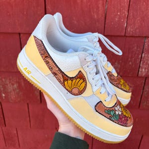 May include: Customized Air Force 1 sneakers with a yellow, white, and brown color scheme. The shoes feature a sunflower design on the Nike swoosh and floral patterns on the toe and heel. The soles are a light brown color.