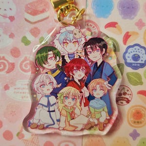 May include: A clear acrylic charm featuring a group of anime-style characters with various hair colors and outfits. The charm has a gold-colored clasp and is attached to a pink background with stickers.