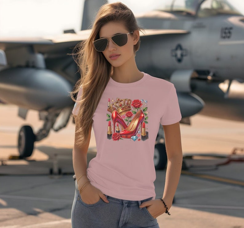 Puede incluir: Una camiseta rosa con un dise&ntilde;o gr&aacute;fico de tacones altos rojos, una corona, pintalabios y una rosa. El dise&ntilde;o est&aacute; centrado en la camiseta.