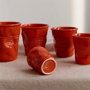 May include: Five orange ceramic mugs with a textured, crumpled design. The mugs are arranged on a light brown linen tablecloth.
