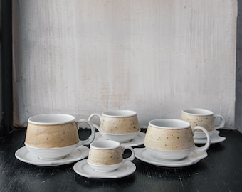May include: A set of five ceramic cups and saucers. The cups have a beige speckled exterior and white interior, with matching white saucers. The cups vary in size, with a large mug and smaller espresso cups. The set is arranged on a dark surface.