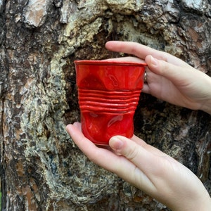Może przedstawiać: Czerwony, pomarszczony kubek metalowy. Kubek jest lekko wgnieciony i ma błyszczące, metaliczne wykończenie.