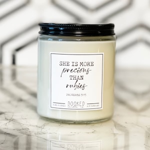 May include: A white candle jar with a black lid and a white label that reads "She is more precious than rubies, Proverbs 3:15, Booked Candles".