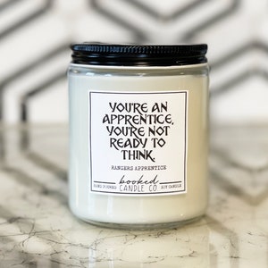 May include: A white soy candle with a black lid and a label that reads "You're an apprentice, you're not ready to think." The label also includes the text "Rangers Apprentice" and "booked candle co."