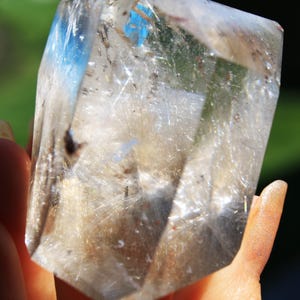May include: A clear quartz crystal with a large amount of inclusions, also known as rutilated quartz. The crystal is faceted and has a point at the top. The crystal is being held in a hand.