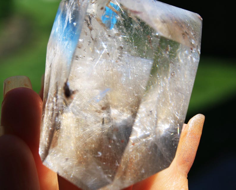 May include: A clear quartz crystal with a large amount of inclusions, also known as rutilated quartz. The crystal is faceted and has a point at the top. The crystal is being held in a hand.